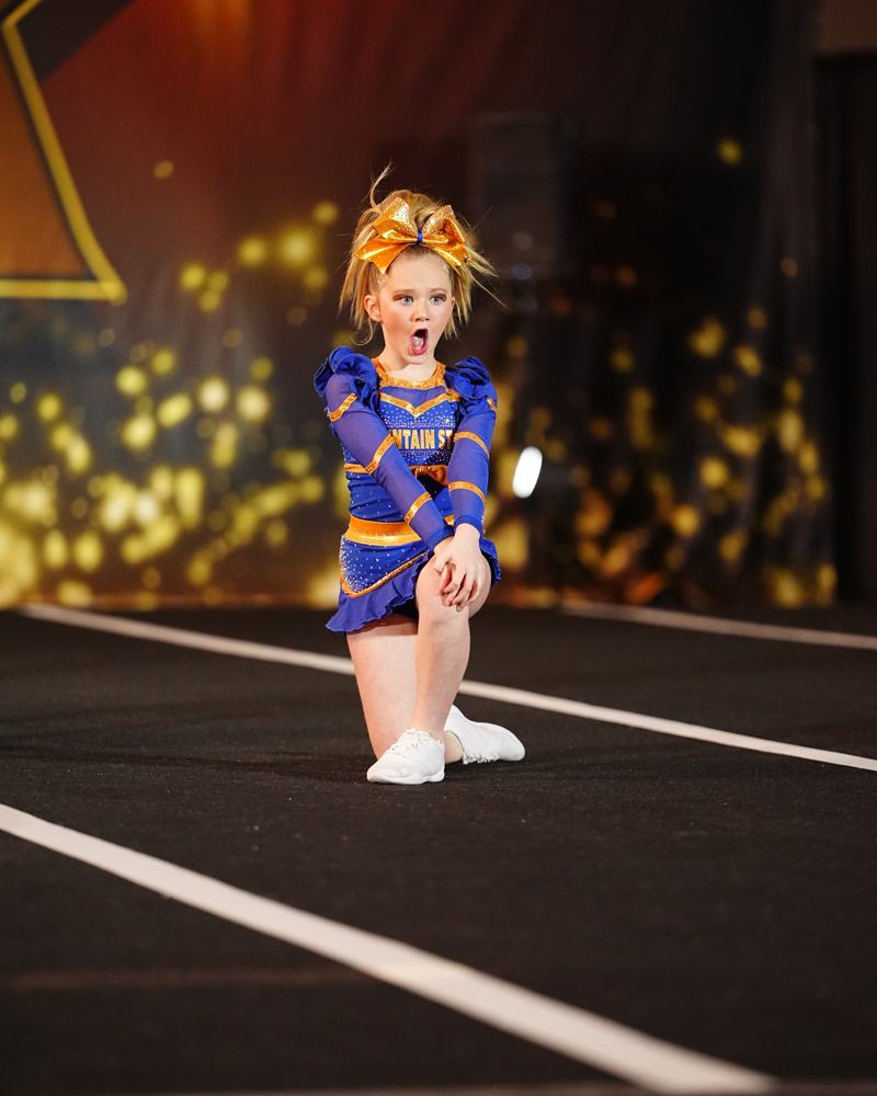 Honeybees athlete in a gold bow hitting a sharp kneeling pose on the competition floor
